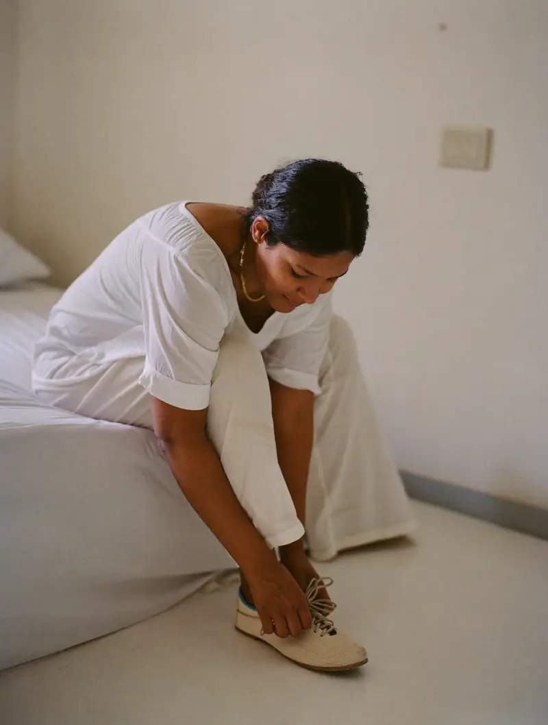 A woman tying her walking shoes on the edge of her bed in morning light.
